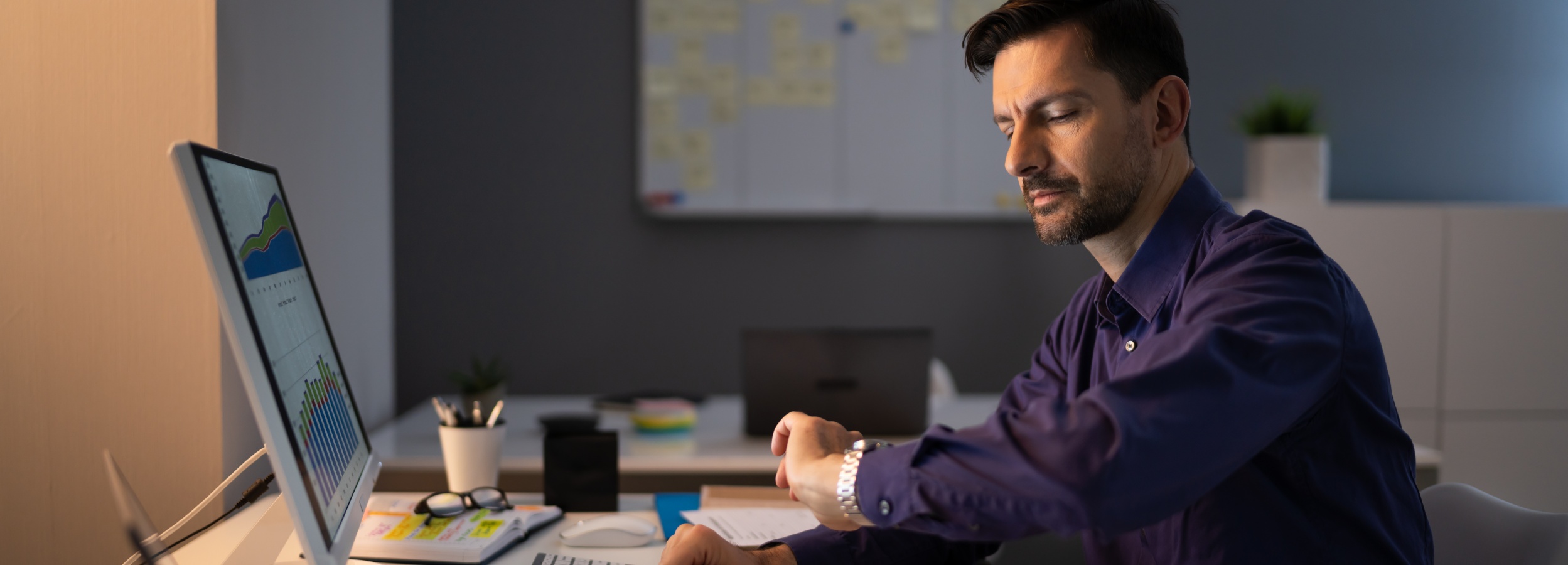 Ein Mann arbeitet spätabends in einem Büro an seinen Computergrafiken und scheint auf seine Uhr zu schauen.

Das Bild zeigt einen Mann mittleren Alters, der in einem dunklen Büro sitzt und aufmerksam auf seinen Computerbildschirm starrt. Er trägt ein dunkelblaues Hemd. An seinem linken Handgelenk trägt er eine silberne Armbanduhr, die er gerade zu betrachten scheint. Sein Gesichtsausdruck ist konzentriert, vielleicht auch leicht erschöpft, was auf lange Arbeitsstunden hindeutet. Der Monitor vor ihm zeigt Diagramme und Grafiken, die auf Datenanalyse oder Finanzberichte hindeuten. Auf dem Schreibtisch liegen ein offenes Notizbuch mit gelben Haftnotizen, eine Brille, eine Computermaus und Tastatur sowie ein Stiftehalter mit einigen Stiften. Im Hintergrund sind weitere Büromöbel und eine Wandtafel mit vielen Notizen zu sehen. Die Beleuchtung ist gedämpft und warm, was auf die späte Stunde oder den Abendbetrieb im Büro schließen lässt. Insgesamt vermittelt das Bild eine Atmosphäre von konzentrierter Arbeit, möglicherweise unter Zeitdruck, und die typische Stimmung eines Büros, das nach Feierabend noch genutzt wird.
