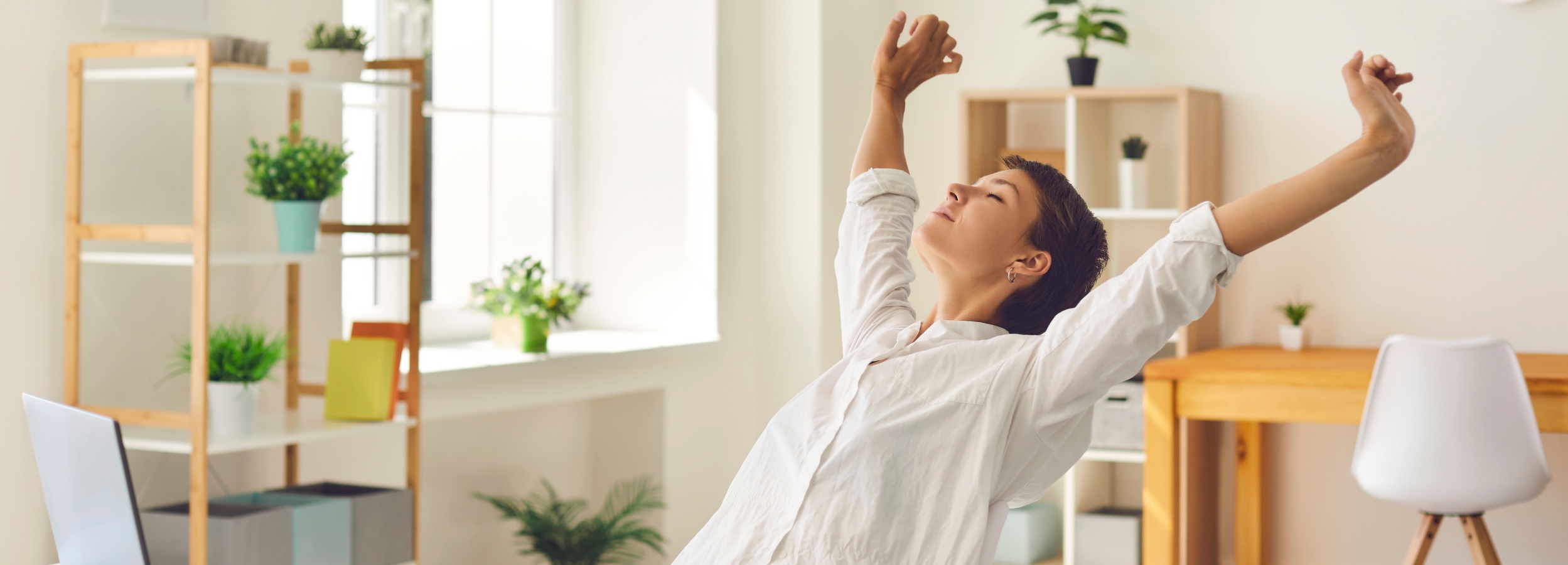 Junge Frau sitzt am Schreibtisch, macht eine Pause von der Arbeit und dehnt steife, angespannte Muskeln