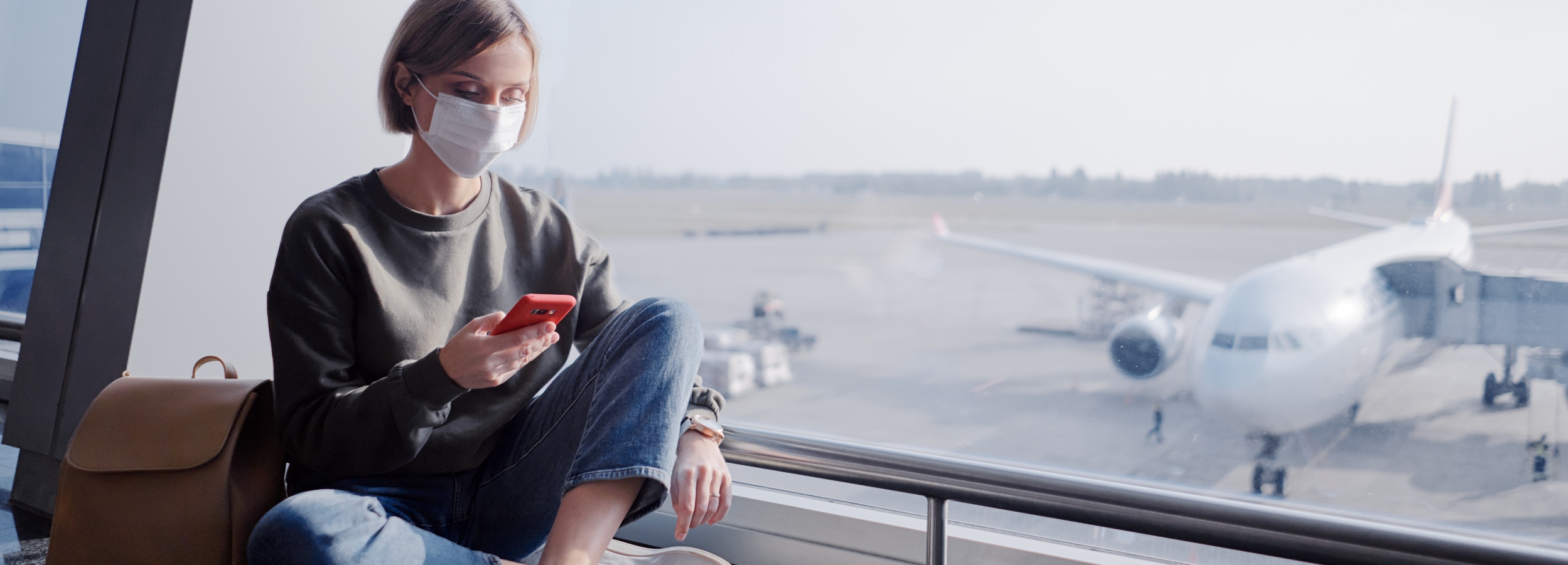 Junge Frau mit weißer Gesichtsmaske auf dem Flughafen schaut auf ihr Handy: Krankschreibung Auslandsurlaub