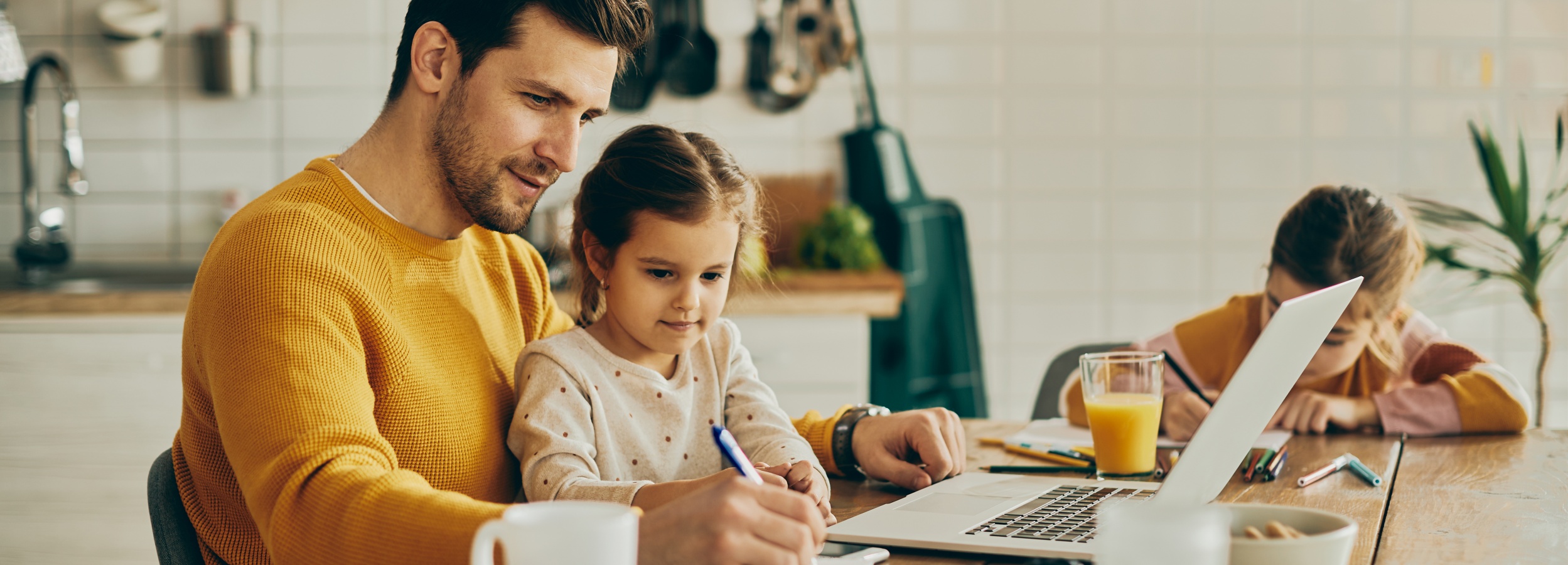 Vater mit zwei Töchtern sitzt mit Laptop und Arbeitsmaterialien am Küchentisch und arbeitet. Ein Kind malt, ein Kind sitzt bei dem Vater auf dem Schoß und schaut ihm bei der Arbeit zu.