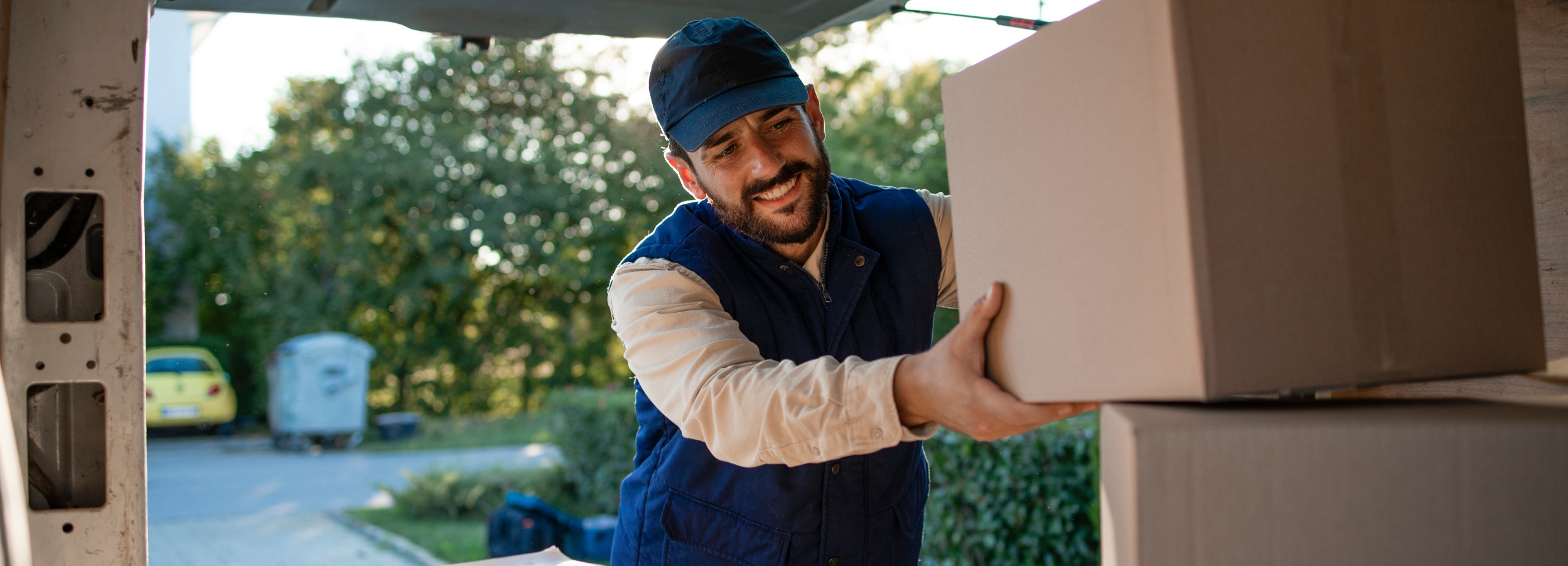 Junger Auslieferungsfahrer mit blauer Schirmmütze und blauer Weste entlädt einen Van mit Paketen.