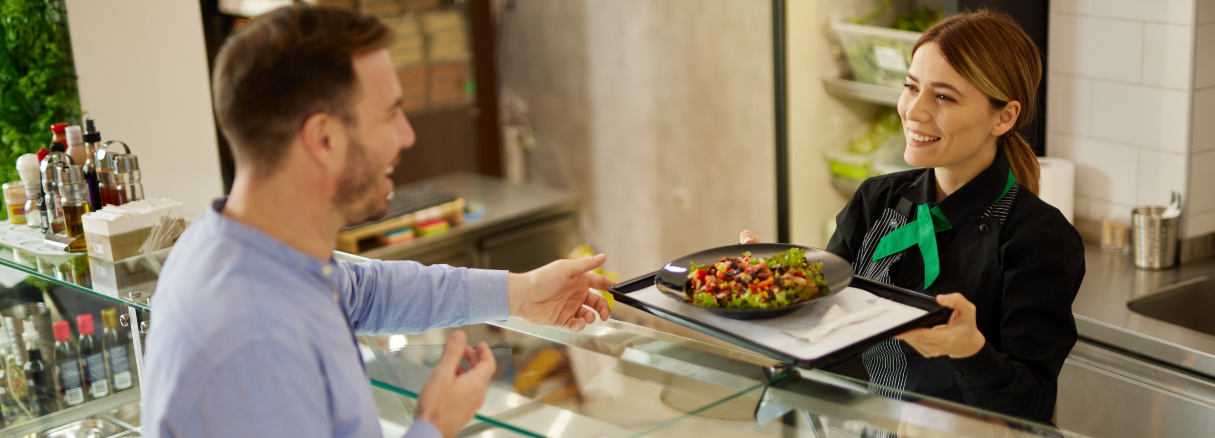 Ein Mann nimmt an der Theke einer Kantine ein Tablett mit einem Salat von einer Mitarbeiterin entgegen.