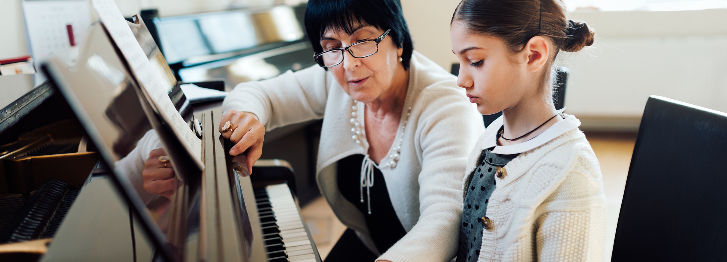 Eine ältere Frau unterrichtet ein junges Mädchen am Klavier.

Auf dem Bild ist eine ältere Frau mit dunklem Haar und Brille zu sehen, die neben einem jungen Mädchen sitzt. Beide sind auf ein Klavier konzentriert, wobei die Frau dem Mädchen offenbar Anweisungen gibt und mit dem Finger auf die Notenblätter zeigt. Das Mädchen, dessen Haar zu einem Dutt hochgesteckt ist und das eine hübsche Strickjacke über einem Kleid mit Herzmuster trägt, blickt ebenfalls auf die Noten. Die Szene spielt in einem hellen Raum, der durch ein Fenster im Hintergrund beleuchtet wird, was eine ruhige und konzentrierte Atmosphäre schafft. Der Fokus liegt auf der Interaktion zwischen Lehrerin und Schülerin, was auf eine liebevolle und geduldige Lernumgebung hindeutet. Die Kleidung der beiden Personen wirkt ordentlich und gepflegt, was die Seriosität der Musikstunde unterstreicht. Die spiegelnde Oberfläche des Klaviers reflektiert die Szene und verleiht dem Bild eine zusätzliche visuelle Ebene.