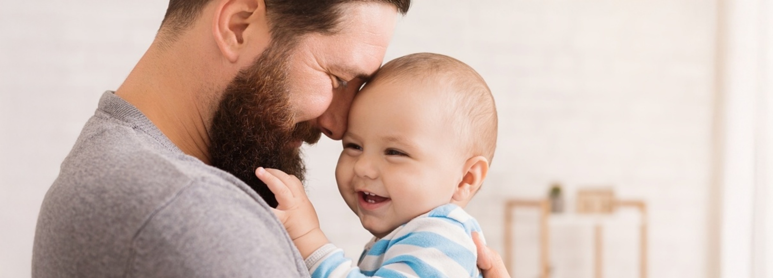 Ein Vater und sein Kind teilen einen intimen und liebevollen Moment der Verbundenheit.

Auf dem Bild ist ein Mann mit einem vollen Bart und einer grauen Strickjacke zu sehen, der sich liebevoll an sein Baby schmiegt. Das Baby, das ein blau-weiß gestreiftes Oberteil trägt, ist ihm zugewandt und strahlt über das ganze Gesicht. Seine Augen sind hell und voller Freude, ein kleiner Mund lacht und zeigt winzige Zähnchen. Die Stirn des Mannes ruht sanft auf der Stirn des Babys, und ein Lächeln zeichnet sich auf seinem Gesicht ab. Die Aufnahme ist eine Nahaufnahme, die sich auf die Intimität und die zarte Bindung zwischen den beiden konzentriert. Der Hintergrund ist unscharf und hell, was die Aufmerksamkeit auf die Personen lenkt und eine warme, friedliche Atmosphäre schafft. Im Hintergrund scheinen verschwommen Möbel und eine kleine Pflanze zu erkennen zu sein, was auf eine häusliche Umgebung hindeutet. Die Stimmung ist überschwänglich, von purer Freude und bedingungsloser Liebe geprägt. Es ist ein Moment der Geborgenheit und des Glücks, der die tiefe emotionale Verbindung zwischen Vater und Kind festhält.