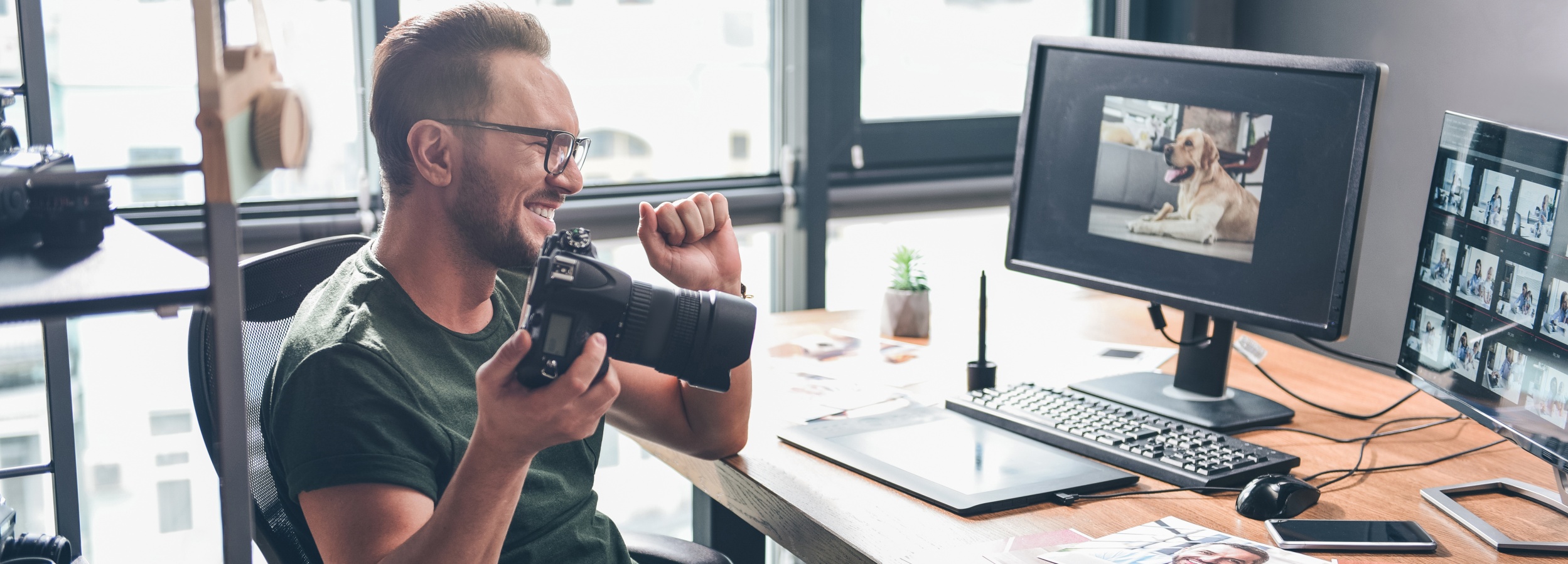 Ein Fotograf feiert einen Erfolg in seinem sonnendurchfluteten Büro, während er seine Ausrüstung und bearbeiteten Ein Fotograf feiert einen Erfolg in seinem sonnendurchfluteten Büro, während er seine Ausrüstung und bearbeiteten Bilder betrachtet.

Das Bild zeigt einen Mann mit Brille und einem dunkelgrünen T-Shirt, der in einem Bürostuhl sitzt und eine Digitalkamera in der rechten Hand hält. Er lächelt breit und hat die linke Faust geballt, was auf Zufriedenheit oder einen Erfolg hindeutet. Vor ihm steht ein Schreibtisch, auf dem zwei Computermonitore, eine Tastatur, eine Maus, ein Grafiktablett und ein kleiner Topfpflanze platziert sind. Der linke Monitor zeigt ein Bild eines liegenden Golden Retrievers. Der rechte Monitor zeigt eine Galerieansicht mit vielen kleinen Vorschaubildern, wahrscheinlich Aufnahmen desselben Motivs oder einer Fotoserie. Auf dem Schreibtisch liegen auch einige ausgedruckte Fotos, darunter eines mit einem lächelnden Mann. Im Hintergrund sind große Fenster zu sehen, durch die Sonnenlicht einfällt und den Raum erhellt. Die Atmosphäre ist hell, positiv und produktiv, was auf die Freude des Mannes an seiner Arbeit und möglicherweise an einem gelungenen Shooting oder einer erfolgreichen Bildbearbeitung hindeutet. Man sieht auch eine Kamera auf einem Regal im Hintergrund.