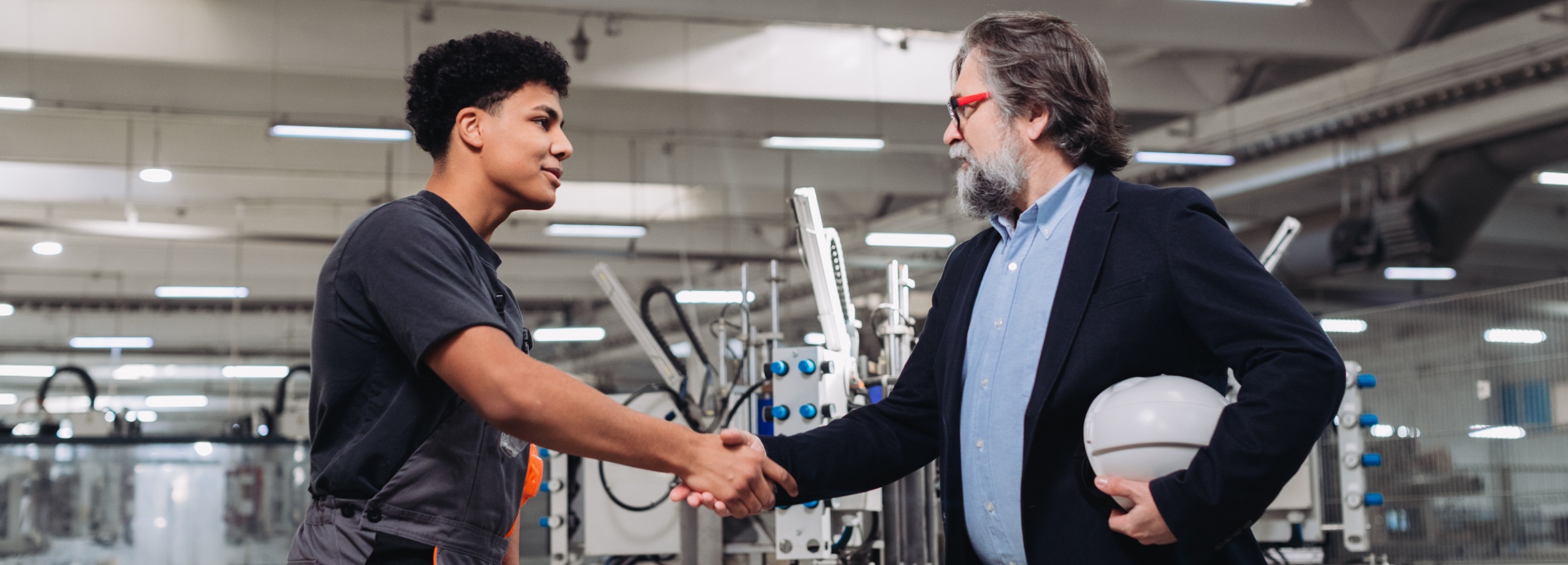 Zwei Männer, einer in Arbeitskleidung und einer in Geschäftskleidung, reichen sich in einer Industriehalle die Hände, was eine geschäftliche Interaktion oder Vereinbarung darstellt.

Auf dem Bild sind zwei Männer abgebildet, die sich die Hände schütteln und im Vordergrund stehen. Der jüngere Mann auf der linken Seite hat dunkle, lockige Haare und trägt ein dunkelgraues T-Shirt mit einer grauen Latzhose, an der ein orangefarbenes Detail zu erkennen ist. Er blickt den älteren Mann an und zeigt ein leichtes Lächeln, das Freundlichkeit oder Zufriedenheit ausdrückt. Der ältere Mann auf der rechten Seite hat graues Haar und einen grauen Bart. Er trägt eine hellblaue Hemd unter einem dunkelblauen Blazer und eine auffällige Brille mit rot-schwarzem Rahmen. In seiner linken Hand hält er einen weißen Schutzhelm. Auch er blickt den jüngeren Mann an und scheint dabei ernst, aber wohlwollend.

Der Hintergrund zeigt eine helle Industrie- oder Fabrikhalle mit hohen Decken. Man erkennt verschiedene Maschinen und Anlagen, die aus Metall zu sein scheinen und mehrere Schläuche, Kabel sowie blaue Bedientasten aufweisen. Die Beleuchtung erfolgt durch mehrere lange, rechteckige Leuchtstoffröhren, die an der Decke montiert sind und für eine gleichmäßige Ausleuchtung sorgen. Es sind keine Tiere im Bild zu sehen.

Die Atmosphäre des Bildes ist professionell und geschäftlich, aber gleichzeitig positiv und kooperativ. Die Geste des Händedrucks vermittelt ein Gefühl von Vertrauen, Vereinbarung oder einer erfolgreichen Zusammenarbeit. Die Stimmung ist konstruktiv und zeigt möglicherweise einen Austausch zwischen einem erfahrenen Manager oder Ingenieur und einem jungen Techniker oder Auszubildenden, was auf Mentoring, eine Einstellung oder einen erfolgreichen Geschäftsabschluss hindeuten könnte.
