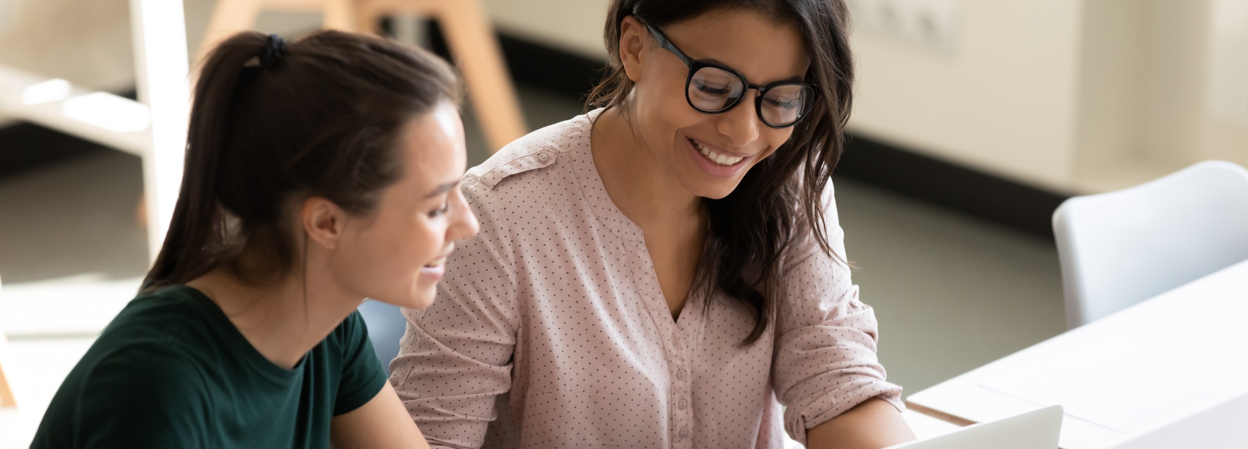 Das Bild zeigt zwei lächelnde Frauen, die gemeinsam an einem Laptop arbeiten und eine positive und kollaborative Arbeitsatmosphäre ausstrahlen.

Im Detail sieht man zwei Frauen, die eng nebeneinander an einem weißen Tisch sitzen. Die Frau links trägt ein dunkelgrünes T-Shirt und hat ihre braunen Haare zu einem Pferdeschwanz gebunden. Sie lacht leicht und schaut auf den Laptop, den die andere Frau hält. Die Frau rechts trägt eine hellrosa Bluse mit kleinen weißen Punkten und eine Brille. Sie lächelt breit und scheint konzentriert auf den Laptop zu sein. Ihr dunkles Haar fällt ihr über die Schultern. Der Laptop ist im Bildausschnitt nur teilweise sichtbar. Im Hintergrund sieht man unscharf die Beine von Stühlen und einen hellen Raum. Die allgemeine Atmosphäre ist freundlich, positiv und konzentriert. Die Stimmung wird durch die lächelnden Gesichter der Frauen und die Nähe zueinander vermittelt. Das Bild strahlt Zusammenarbeit und positive Arbeitsbeziehungen aus.