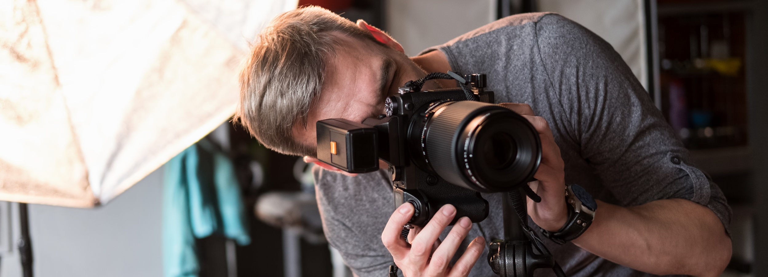 Das Bild zeigt einen konzentrierten Fotografen, der durch eine professionelle Kamera hindurch in die Linse blickt.

Im Detail zeigt das Bild einen Mann mittleren Alters mit kurzen, grau melierten Haaren. Er trägt ein schlichtes, graues T-Shirt und eine Armbanduhr mit Lederarmband. Sein Gesichtsausdruck ist konzentriert und ernst; er ist ganz auf das Fotografieren fokussiert. Er hält eine große, professionelle Spiegelreflexkamera mit einem langen Teleobjektiv. An der Kamera ist ein externes Blitzgerät angebracht. Die Kamera steht auf einem Stativ. Im Hintergrund ist unscharf ein Fotostudio mit Beleuchtungsequipment (ein großer, weicher Diffusor ist teilweise sichtbar) und weitere, nicht genau erkennbare Gegenstände zu sehen. Die Atmosphäre ist ruhig und konzentriert, fast etwas düster, aufgrund der gedämpften Beleuchtung. Die Stimmung ist professionell und intensiv, die Konzentration des Fotografen steht im Vordergrund.