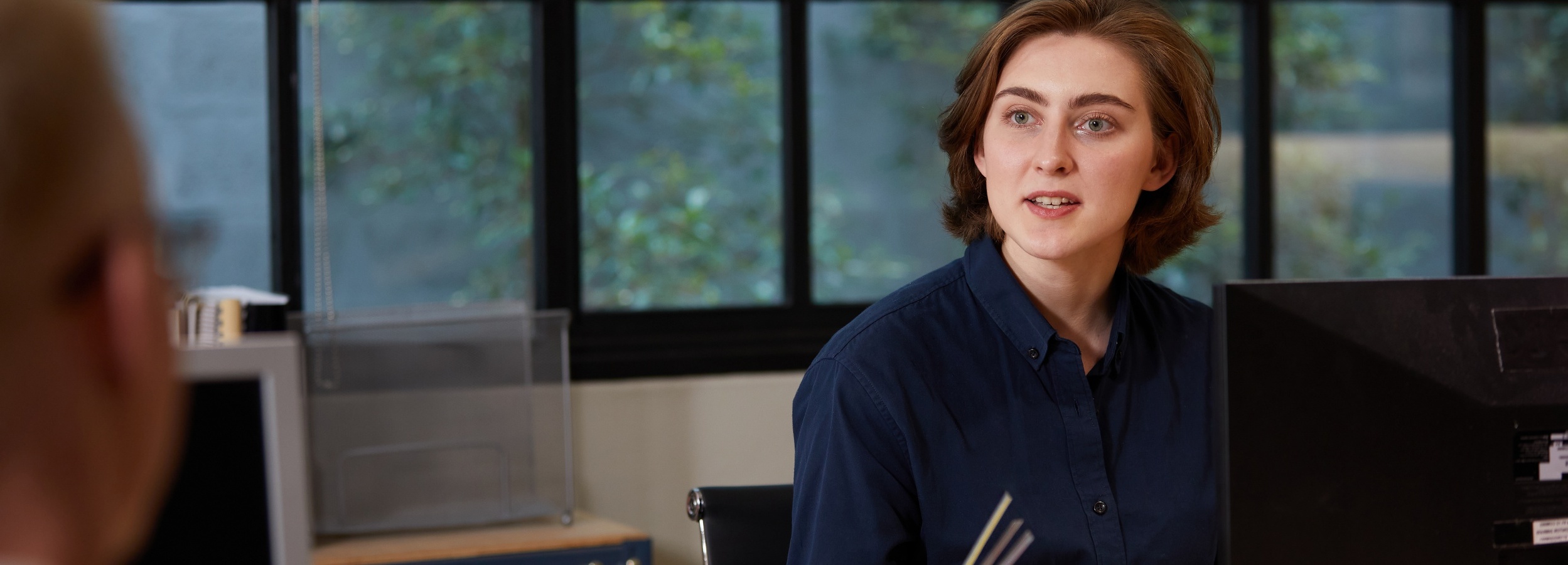 Das Bild zeigt eine junge Frau in einem Büro, die mit einem Kollegen spricht.

Im Detail zeigt das Bild den Oberkörper einer jungen Frau mit hellbraunen, schulterlangen Haaren, die ein dunkelblaues Hemd trägt. Sie sitzt an einem Schreibtisch, vor dem ein schwarzer Computermonitor steht. Ihr Blick ist nach rechts gewandt, wo man nur den Hinterkopf einer anderen blondhaarigen Person sieht, was darauf hindeutet, dass sie sich in einem Gespräch befindet. Im Hintergrund ist ein Fenster zu sehen, das einen Blick ins Grüne freigibt, was dem Raum eine beruhigende Atmosphäre verleiht. Auf der rechten Seite des Bildes sind Teile eines weiteren Schreibtisches mit Bürobedarf zu erkennen. Die Atmosphäre wirkt professionell und gleichzeitig entspannt, was durch das natürliche Licht und die Pflanzen im Hintergrund verstärkt wird.