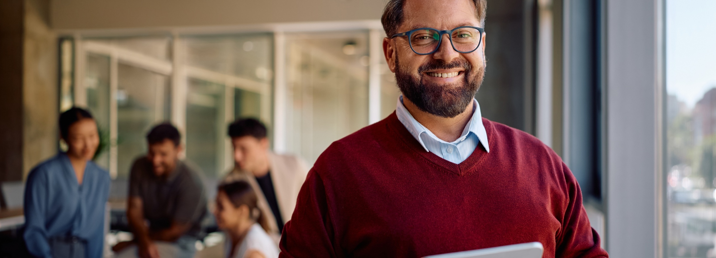 Ein Mann mit Brille lächelt in die Kamera, während er im Hintergrund ein unscharfes Teammeeting leitet.

Das Bild zeigt einen Mann mit Bart und Brille, der freundlich lächelt und ein Tablet hält, während im Hintergrund eine Gruppe von vier Personen in einem Meetingraum zu sehen ist. Der Mann trägt einen roten Pullover über einem hellblauen Hemd. Im Hintergrund sitzen oder stehen die Teammitglieder und scheinen in ein Gespräch vertieft zu sein. Die Atmosphäre wirkt professionell und entspannt zugleich, mit einem Fokus auf Zusammenarbeit und positiver Kommunikation. Das Licht ist hell und natürlich, was die offene und einladende Stimmung des Raumes unterstreicht.