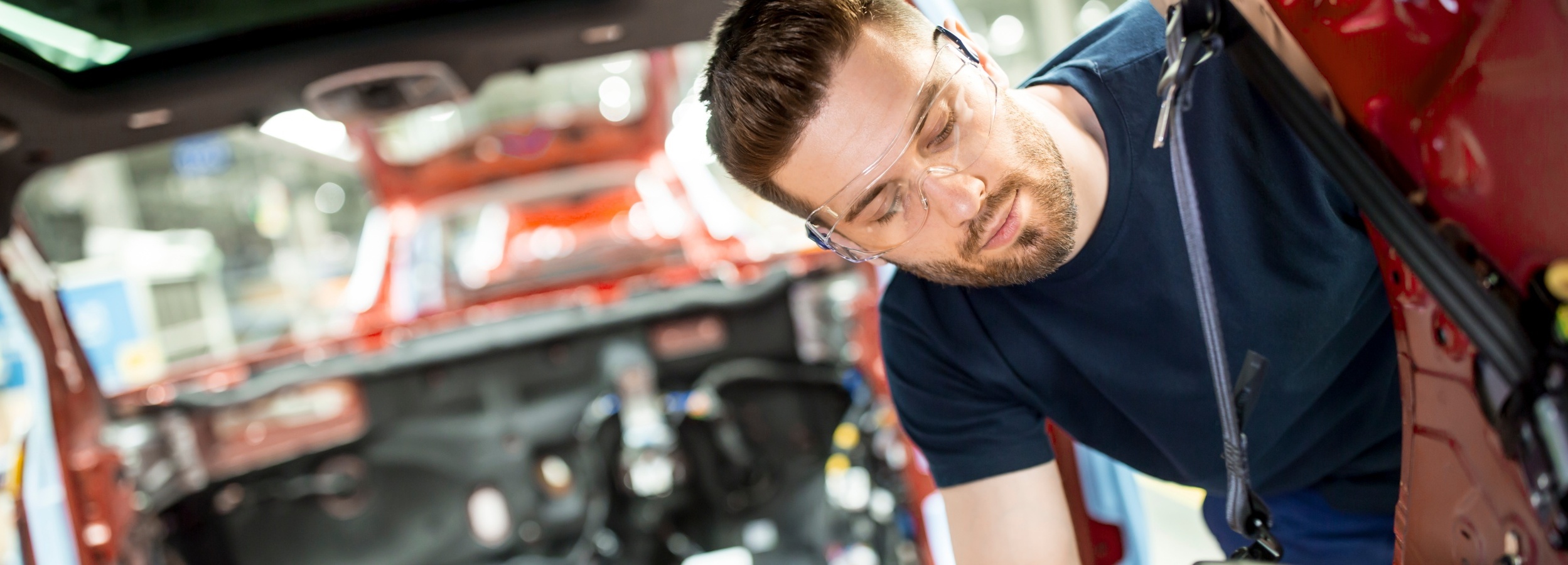 Ein Mann mit Schutzbrille arbeitet konzentriert an der Innenausstattung eines roten Autos. 

Das Bild zeigt einen Mann mit Bart und kurzen braunen Haaren, der eine Schutzbrille trägt und in ein rotes Auto hineinlehnt. Er scheint an der Inneneinrichtung oder Verkabelung des Fahrzeugs zu arbeiten. Sein Gesicht ist nach unten gerichtet, und seine Augen sind auf die Arbeit konzentriert. Er trägt ein dunkelblaues T-Shirt.

Der Hintergrund zeigt das Innere des Autos, das in leuchtendem Rot lackiert ist, sowie verschiedene Kabel und mechanische Teile, die zugänglich sind. Das Sonnenlicht scheint von oben durch ein Schiebedach und beleuchtet den Innenraum. Die Atmosphäre ist hell und professionell, was auf eine Werkstatt oder Produktionsstätte hindeuten könnte.