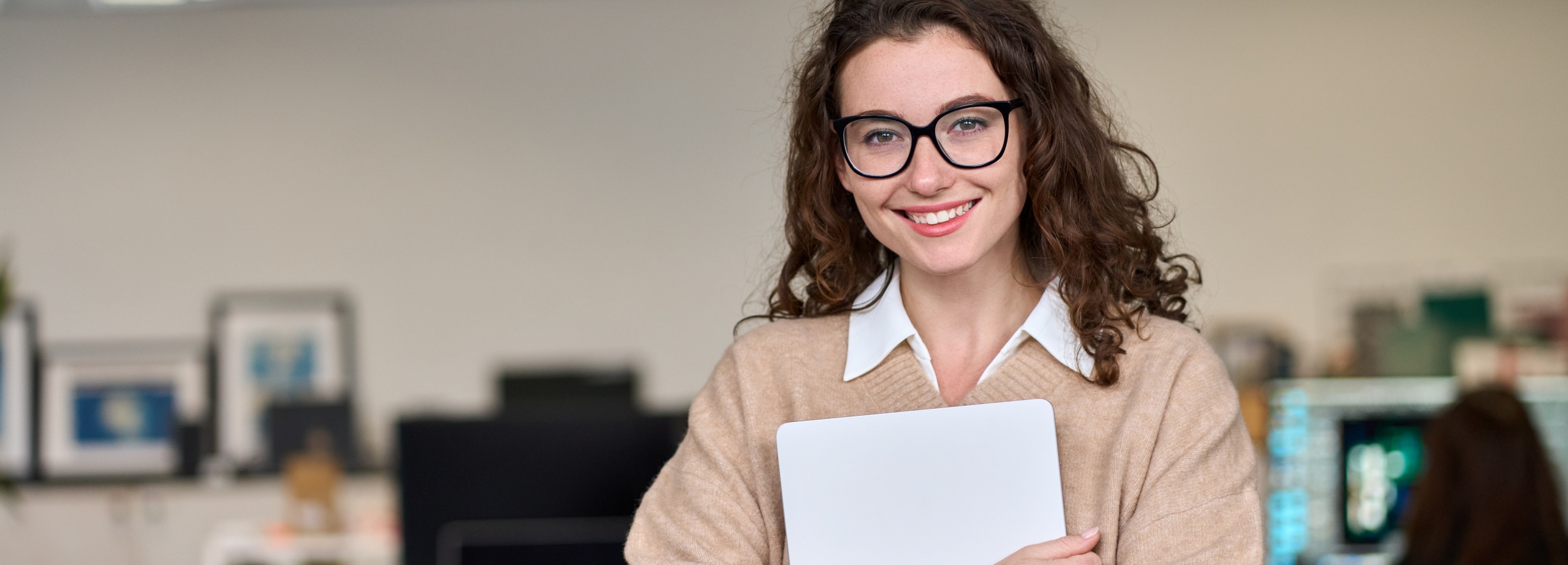 Das Bild zeigt eine lächelnde junge Frau mit Laptop vor einem Bürohintergrund.

Im Detail ist eine junge Frau mit dunklen, lockigen Haaren und Brille zu sehen, die direkt in die Kamera lächelt. Sie trägt eine weiße Bluse unter einem beigen Strickpullover. In ihren Händen hält sie einen weißen Laptop. Im Hintergrund ist ein unscharfer Büroraum erkennbar. Man sieht Schreibtische, Monitore und an der Wand hängende Bilder. Die allgemeine Atmosphäre wirkt professionell und einladend, die Stimmung ist freundlich und positiv.