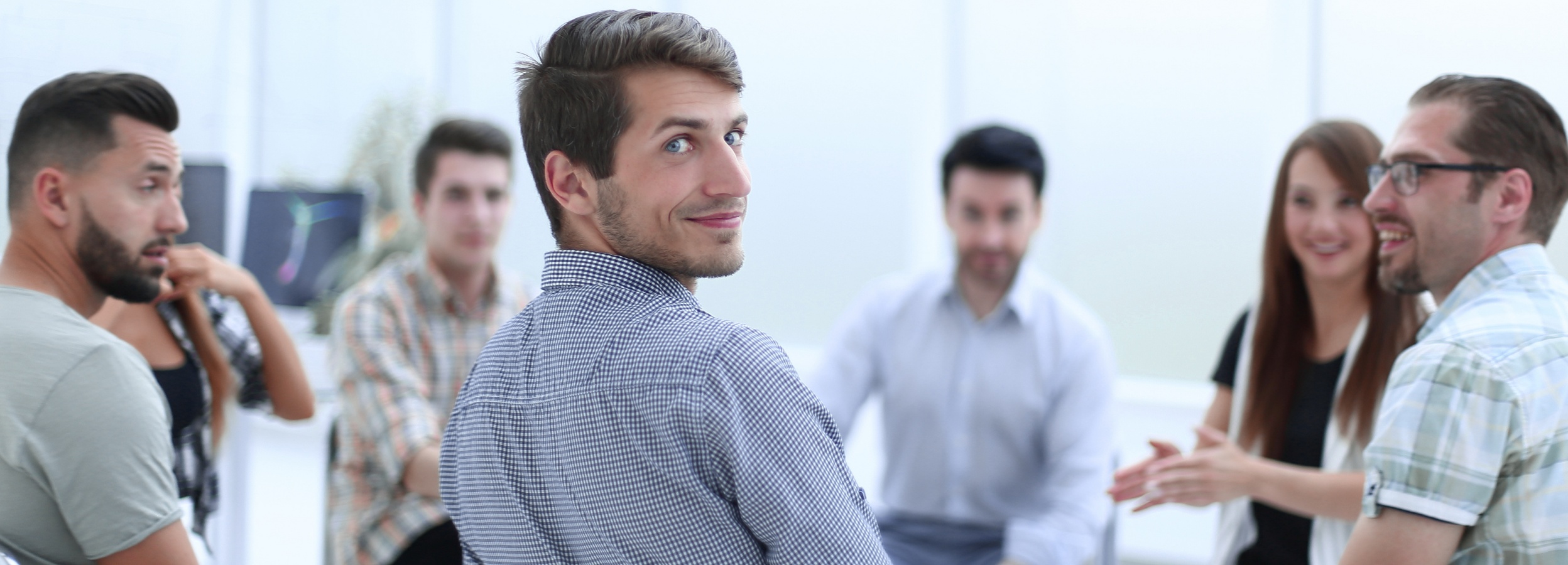 Das Bild zeigt eine Gruppe von Menschen in einem Büro, wobei ein Mann im Vordergrund hervorsticht, der über seine Schulter lächelt.

Im Detail zeigt das Bild eine Gruppe von etwa sieben Personen, die sich in einem hellen, modernen Büroumfeld befinden. Der Fokus liegt auf einem Mann im Vordergrund, der ein blau-weiß kariertes Hemd trägt und direkt in die Kamera lächelt, während er über seine Schulter schaut. Er hat dunkles Haar und blaue Augen, und sein Gesichtsausdruck wirkt freundlich und einladend.

Um ihn herum befindet sich der Rest der Gruppe, die sich scheinbar in einer Diskussion oder einem Meeting befindet. Die Personen sind teilweise unscharf, was darauf hindeutet, dass der Fokus auf dem Mann im Vordergrund liegt. Sie tragen verschiedene Arten von Kleidung, darunter Hemden, Blusen und T-Shirts. Einige von ihnen lächeln oder scheinen aktiv an der Unterhaltung teilzunehmen. Im Hintergrund sind Büromöbel und Computer zu erkennen, was den Eindruck eines geschäftigen Arbeitsplatzes verstärkt.

Die Atmosphäre des Bildes wirkt positiv und dynamisch, was durch die lächelnden Gesichter und die offene Körpersprache der Personen unterstrichen wird. Die Stimmung ist entspannt und kooperativ, was auf ein gutes Arbeitsklima hindeutet.